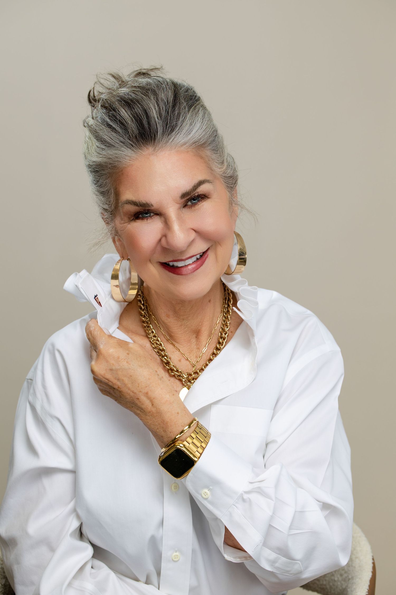 Elegant older woman with gray hair wearing white shirt, gold jewelry, and smartwatch, smiling confidently.