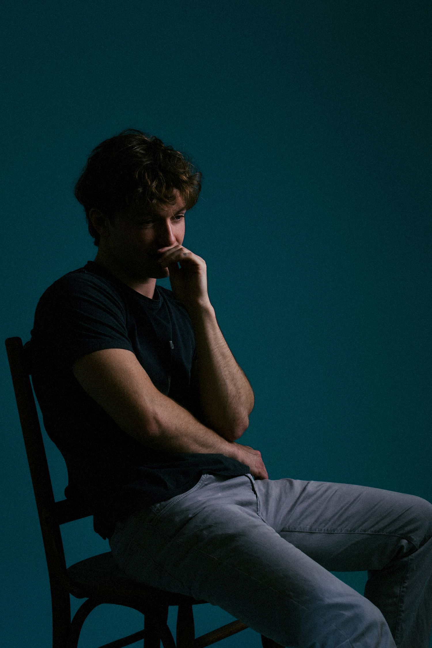 Pensive young man sitting on chair in dark teal studio, resting chin on hand in thoughtful pose.