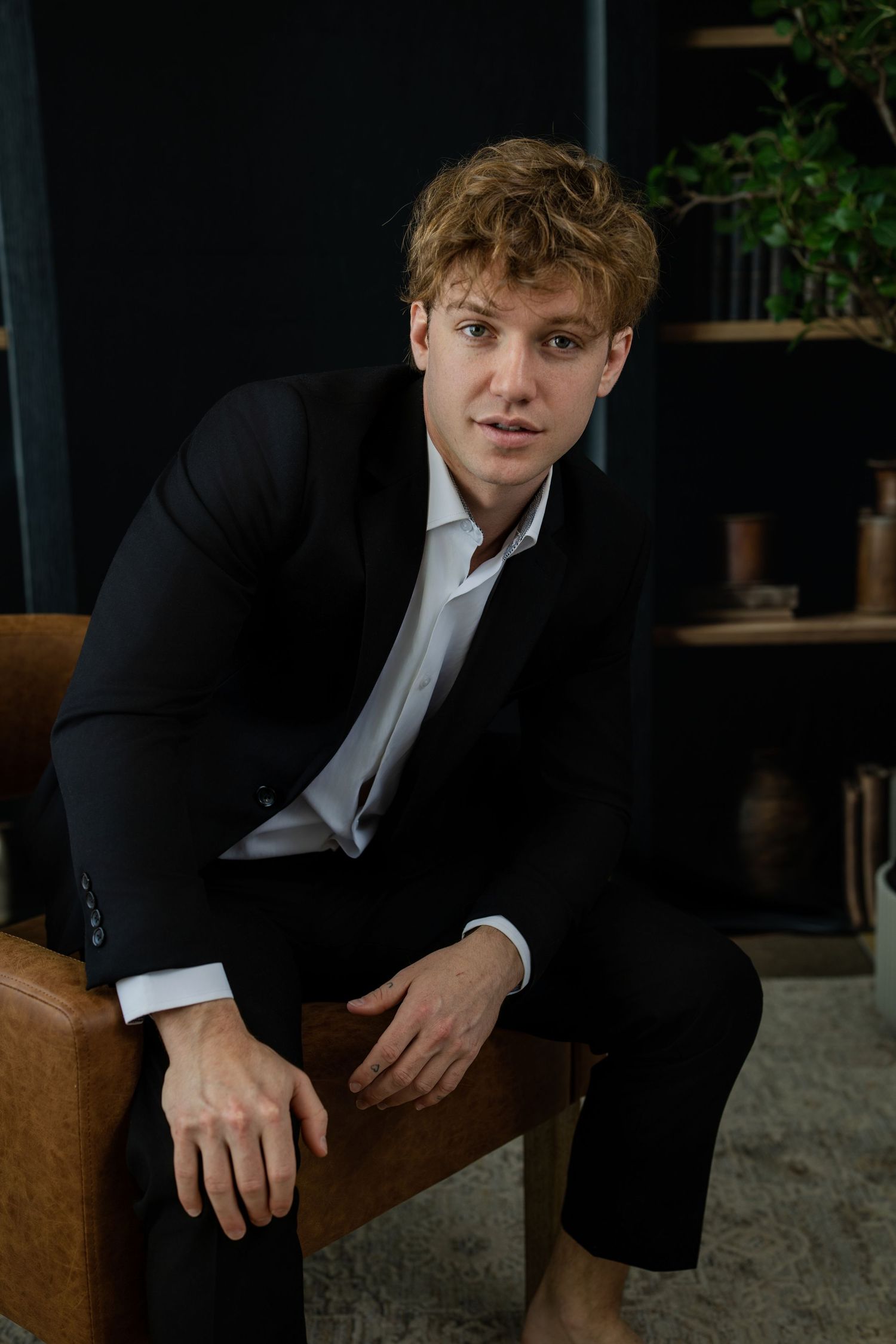 Young man with curly red hair in black suit sitting on leather chair in modern dark interior room.