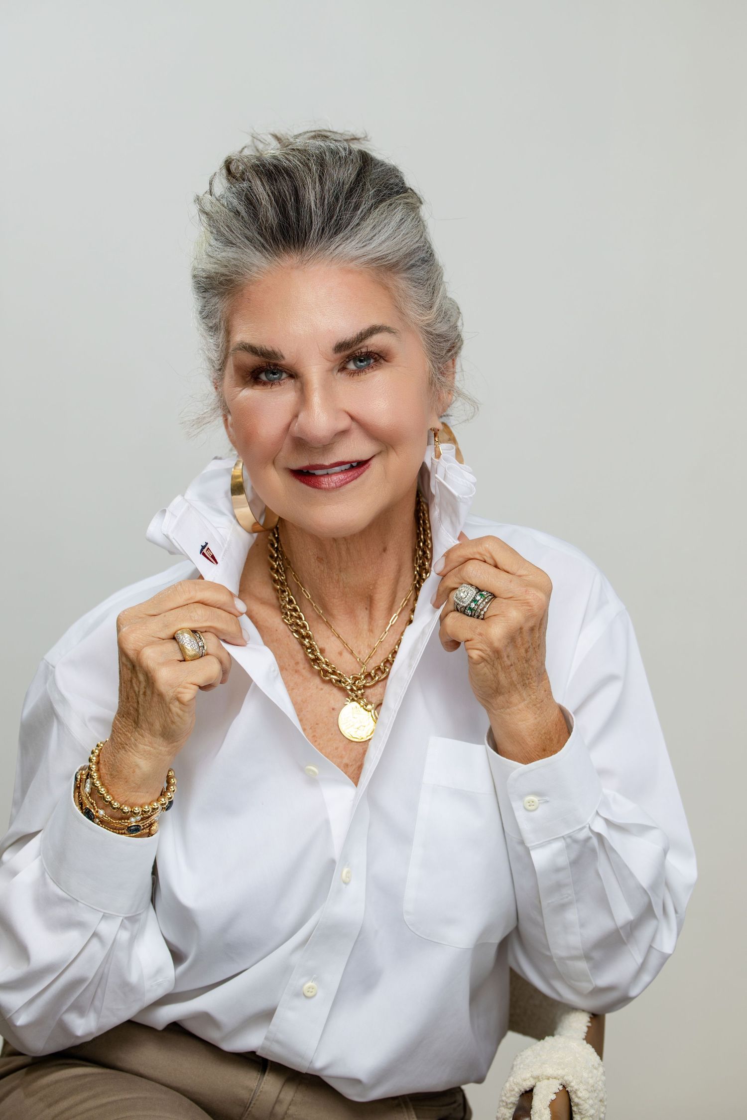 Elegant older woman with gray hair wearing white shirt and gold jewelry, smiling confidently in a studio portrait.