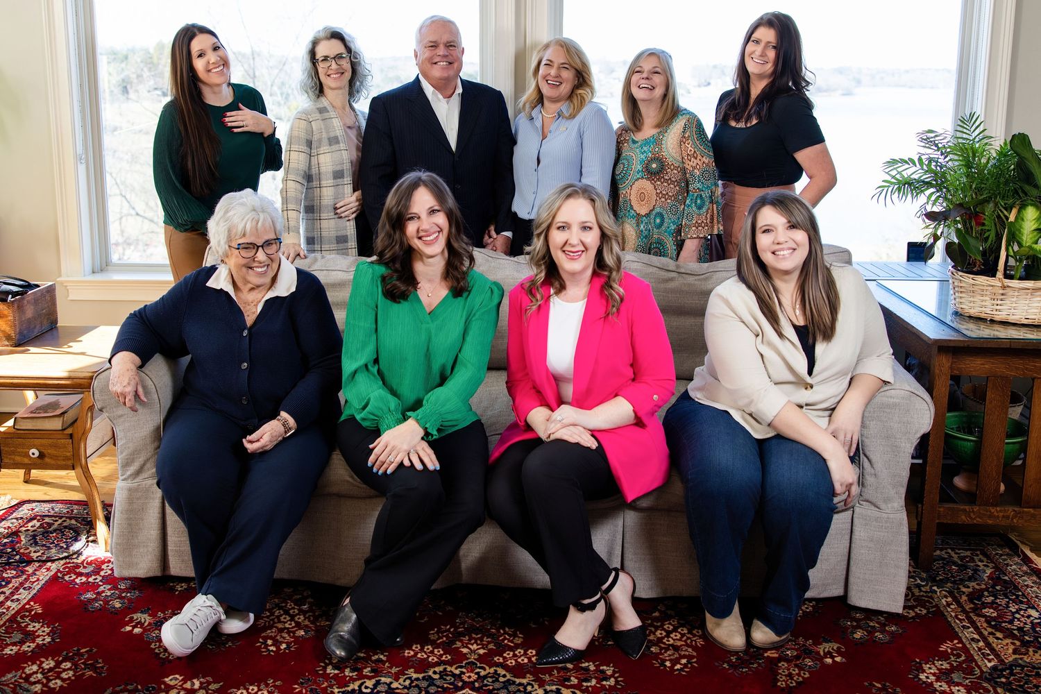 Professional group photo of ten smiling women posed together in a warmly decorated living room setting.