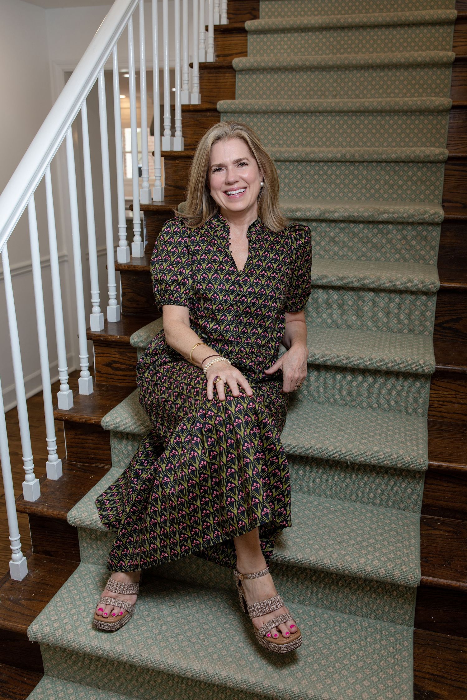 Smiling woman in patterned dress sitting on carpeted staircase with wooden steps and white balusters.