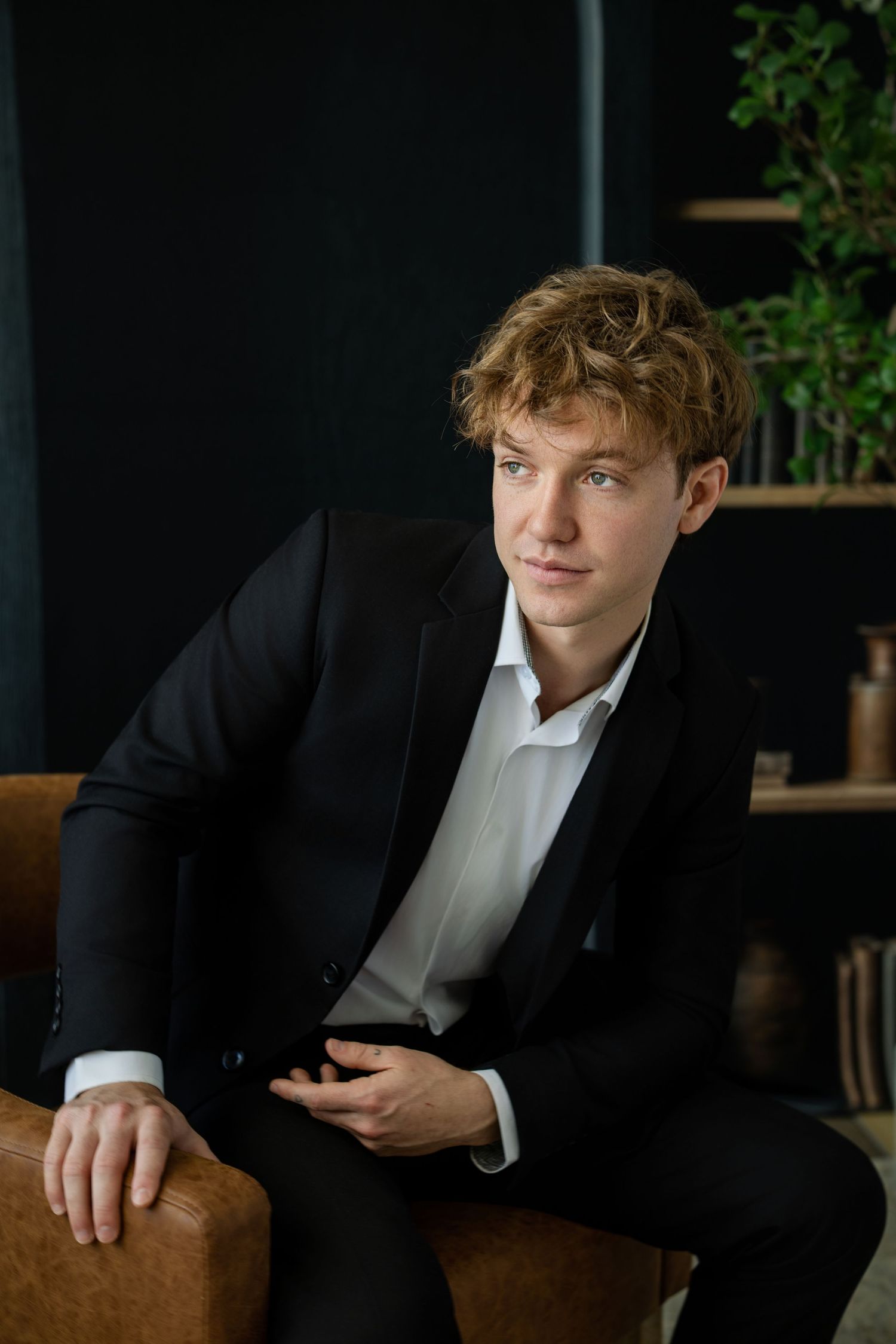 Young man in black suit and white shirt sitting on leather chair holding smartphone in modern office.