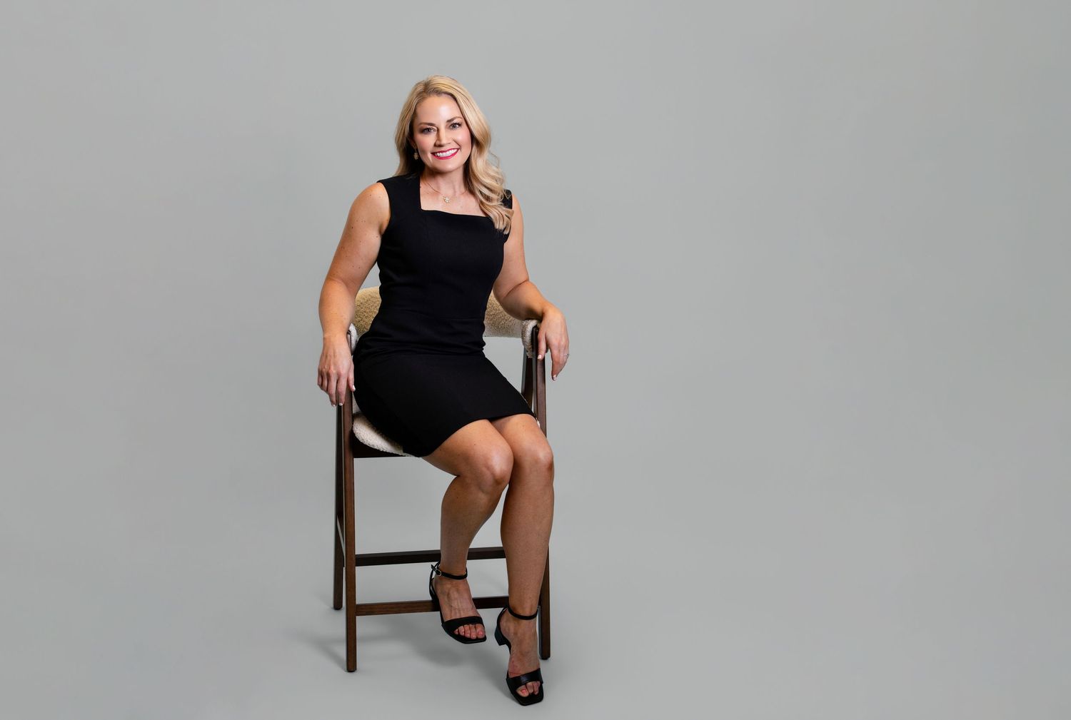 Smiling blonde woman in black dress seated on wooden chair against gray background in professional studio photo.