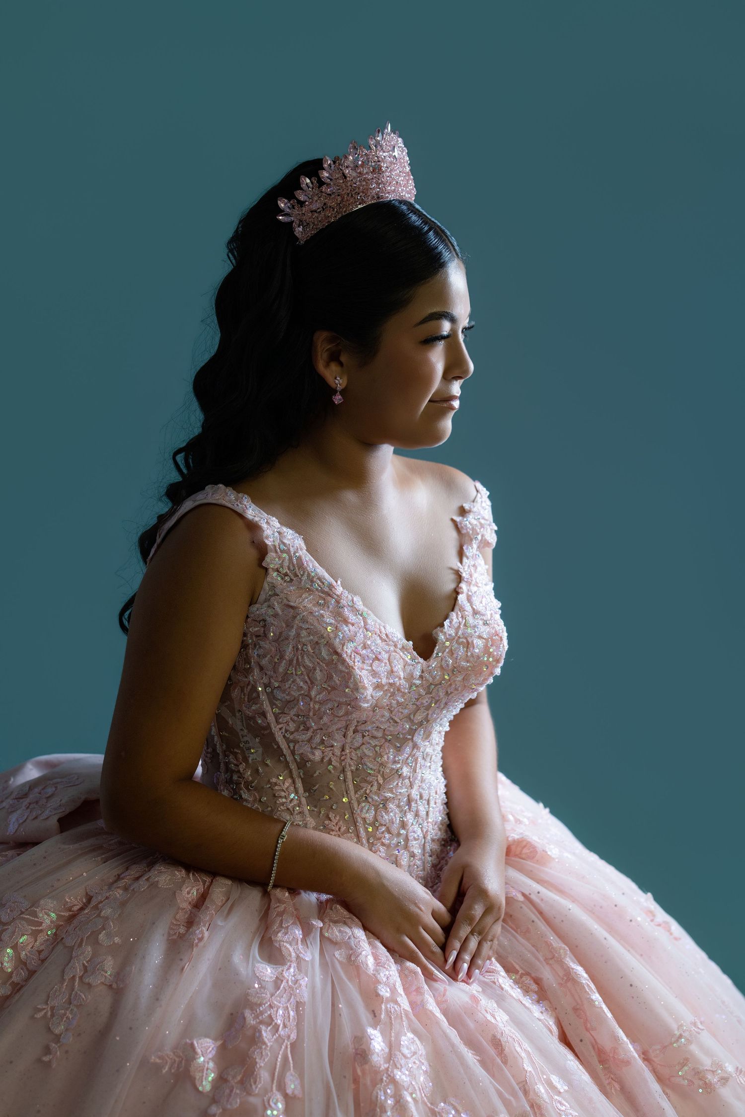 Young Latina woman in pink quinceañera ball gown with floral lace and tiara poses gracefully against teal background.