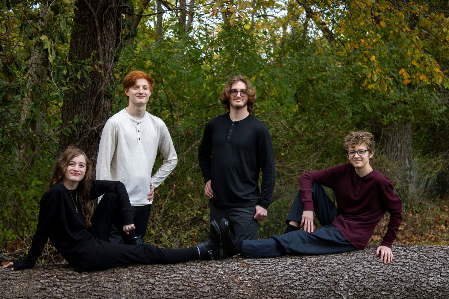 Four young adults pose outdoors on a fallen log surrounded by autumn trees in a casual group portrait.