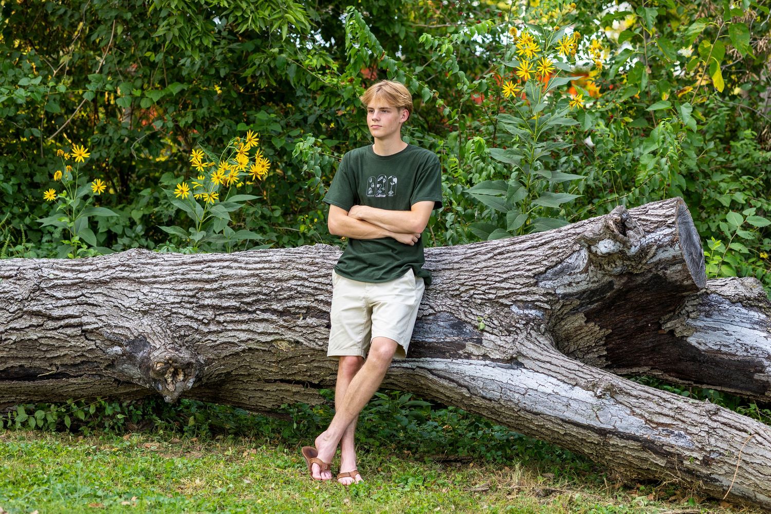 Teen boy in green t-shirt and khaki shorts leaning against a large fallen log in a lush green park.