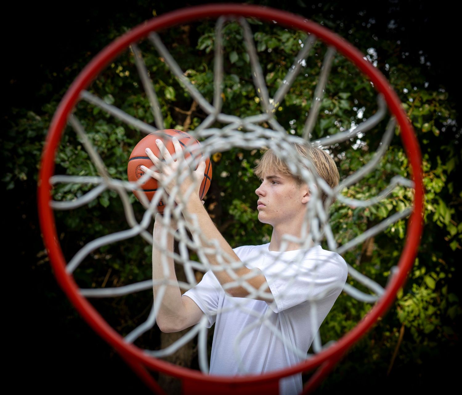 Young male basketball player shooting through an outdoor hoop, viewed from below with green trees in background.