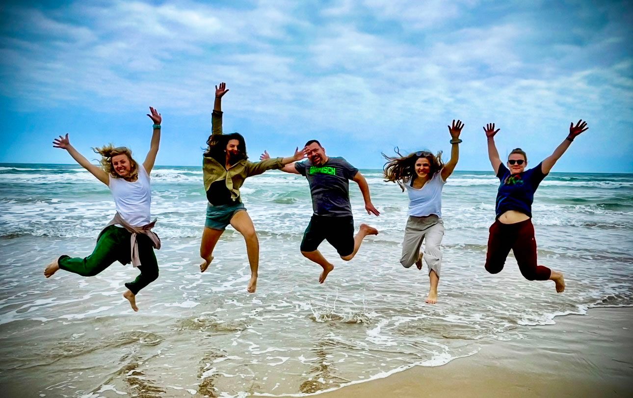 Five people jumping in unison on a beach with waves and cloudy sky in the background.