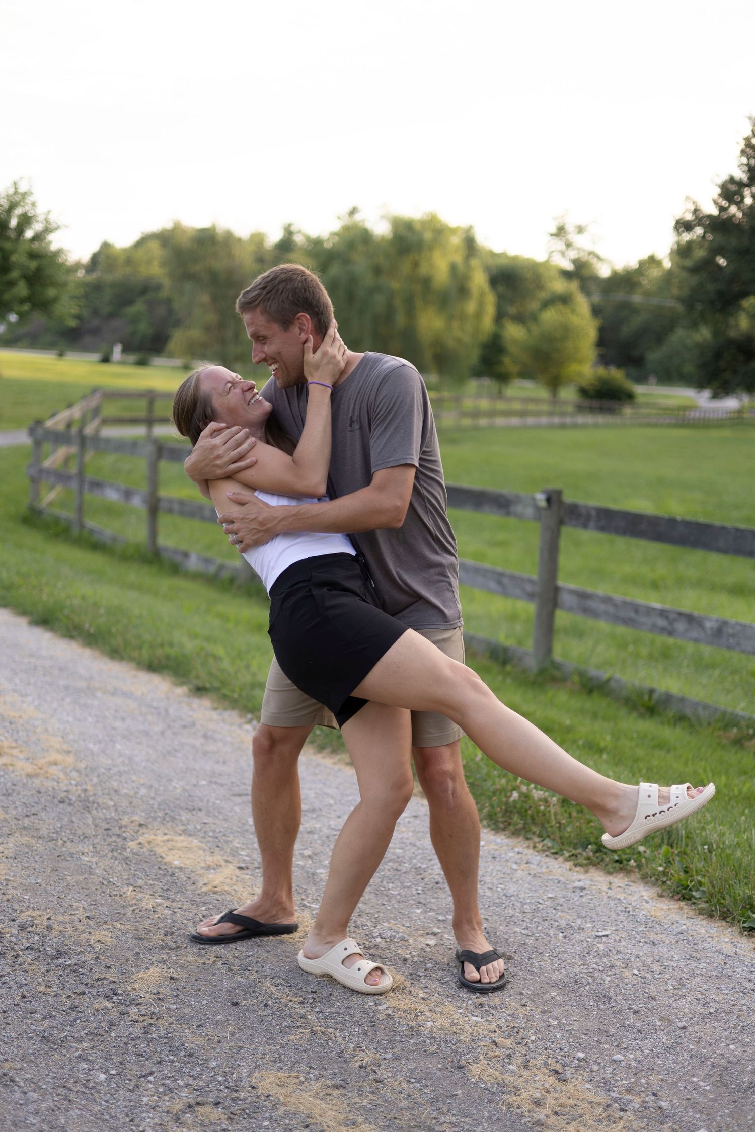 A young couple embracing and laughing together on a rural gravel path surrounded by green fields and wooden fencing.