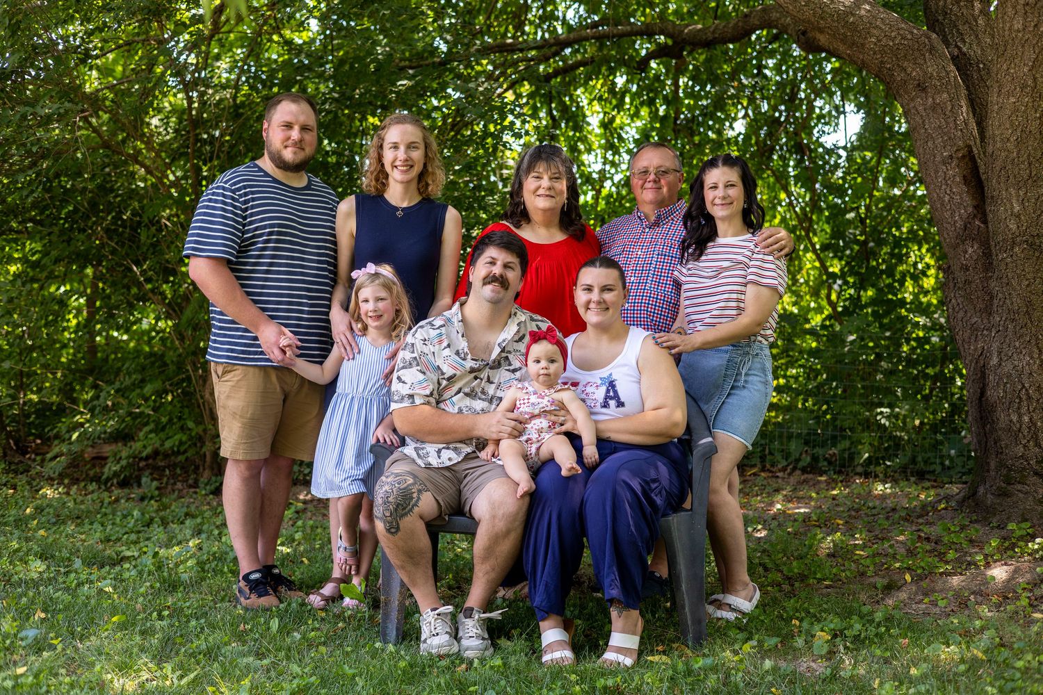 Large family group portrait outdoors under a tree, featuring adults and children smiling in casual attire.