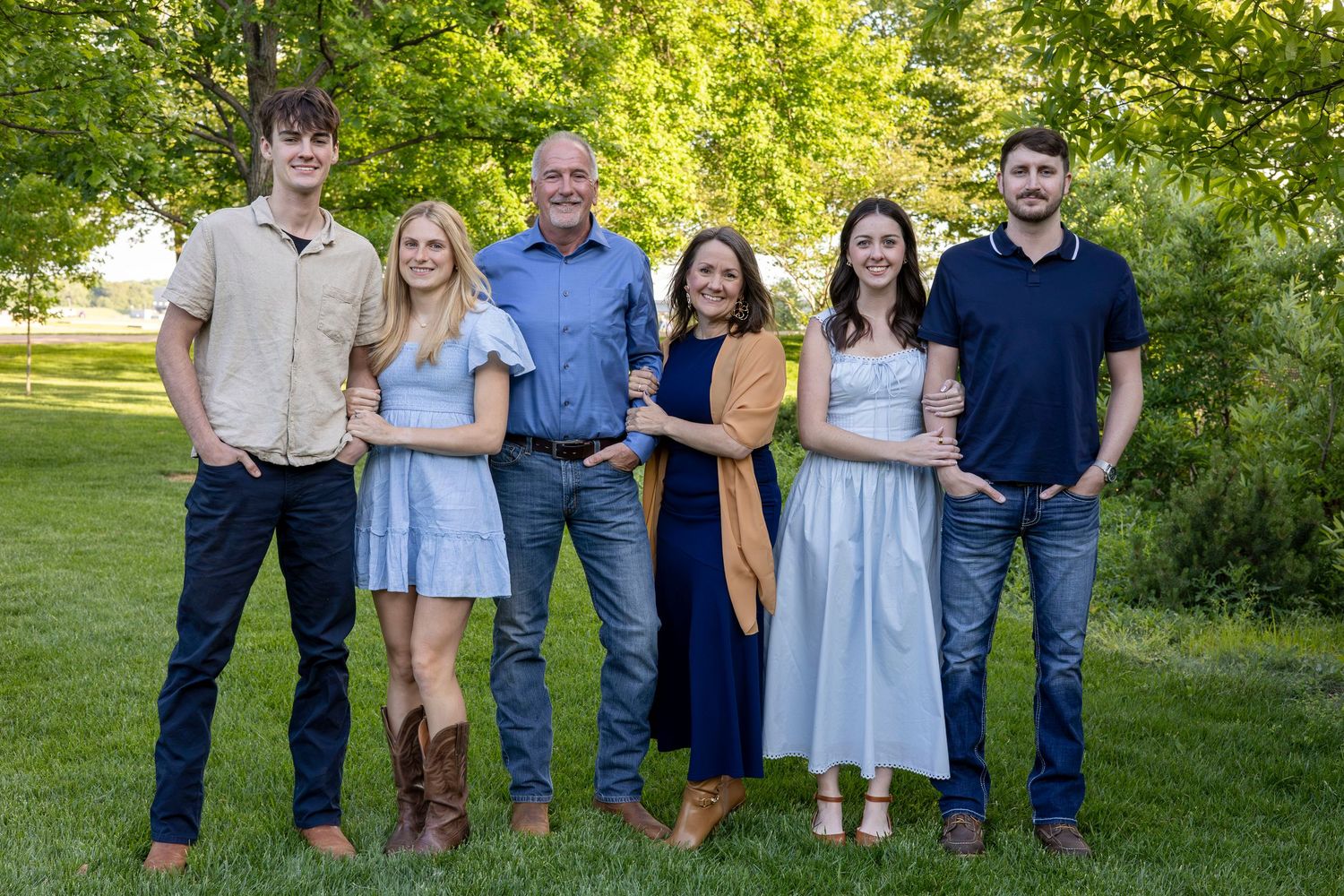 Family of six posing outdoors on green grass surrounded by lush trees in a warm, sunny family portrait.