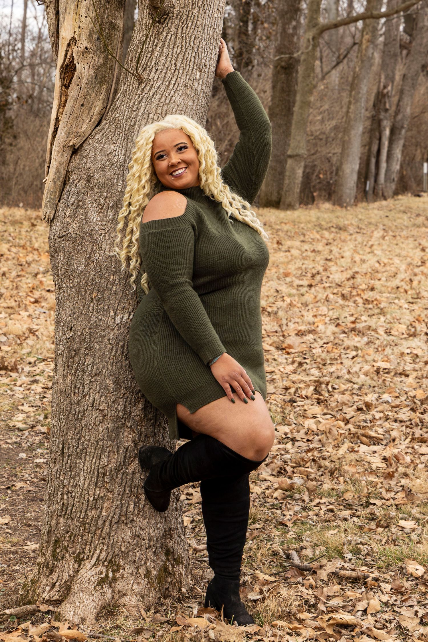 A pregnant woman in a green sweater poses against a tree trunk in a forest filled with fallen autumn leaves.