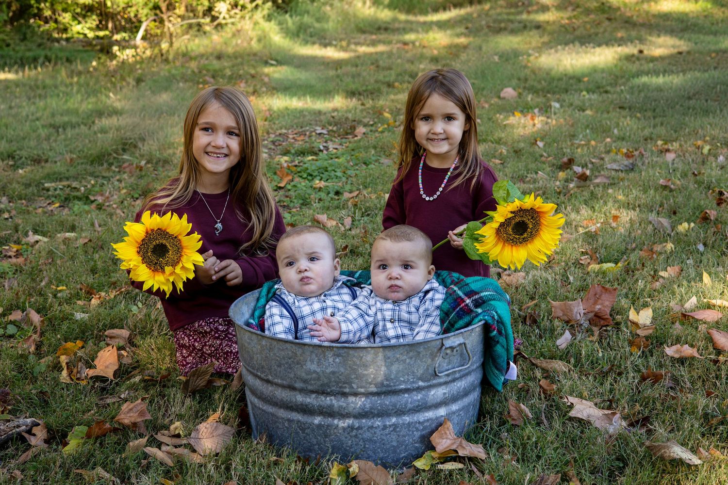 Two girls holding sunflowers sit behind two baby twins in a metal tub on a grassy lawn covered in autumn leaves.