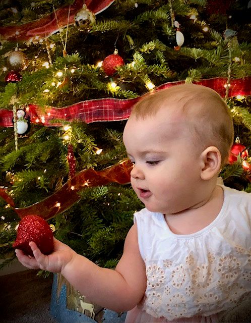 A small child in white dress examines a red ornament in front of a lit Christmas tree.
