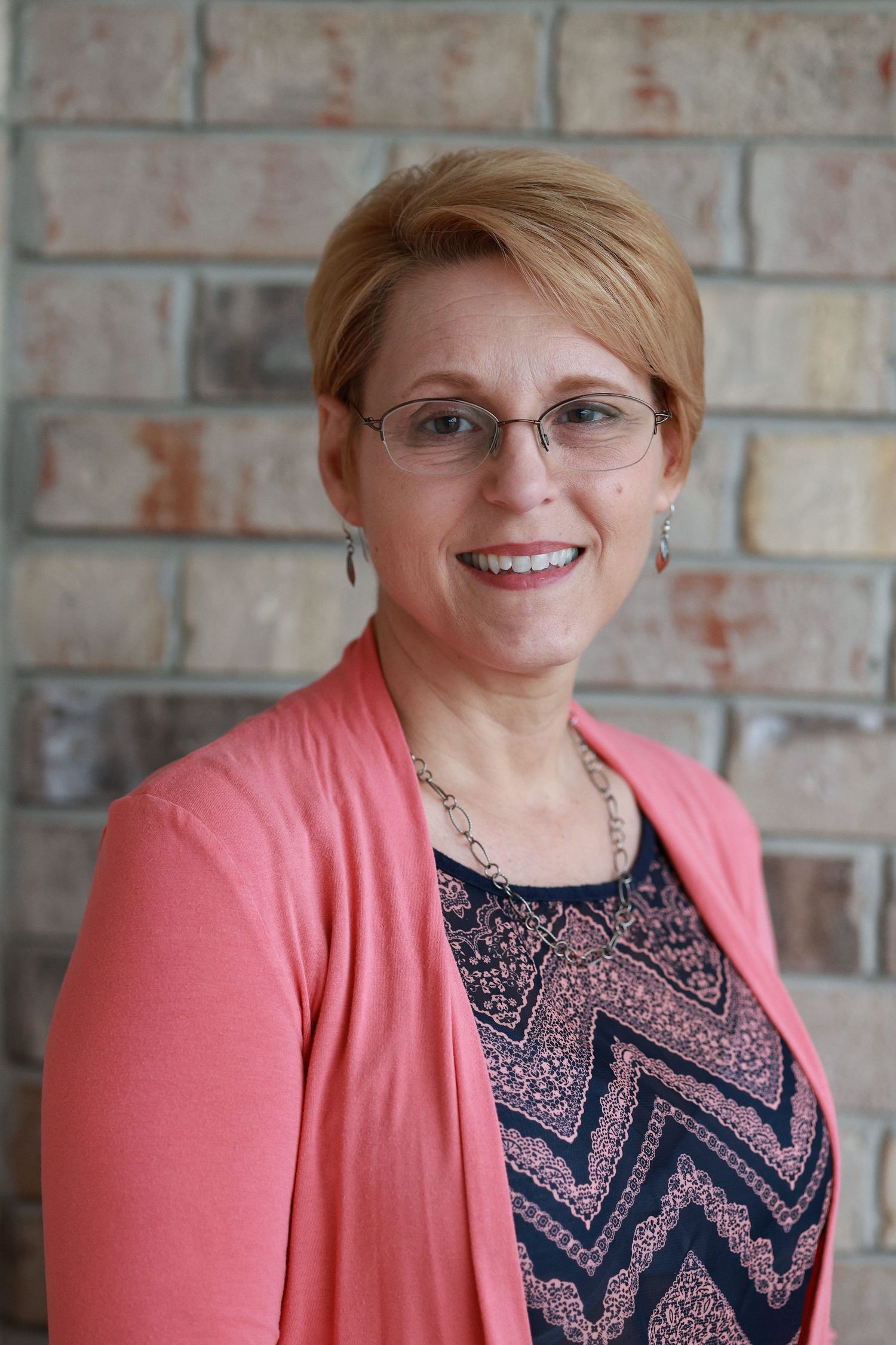 Smiling woman with short hair and glasses wearing a pink cardigan and patterned top in front of a brick wall.
