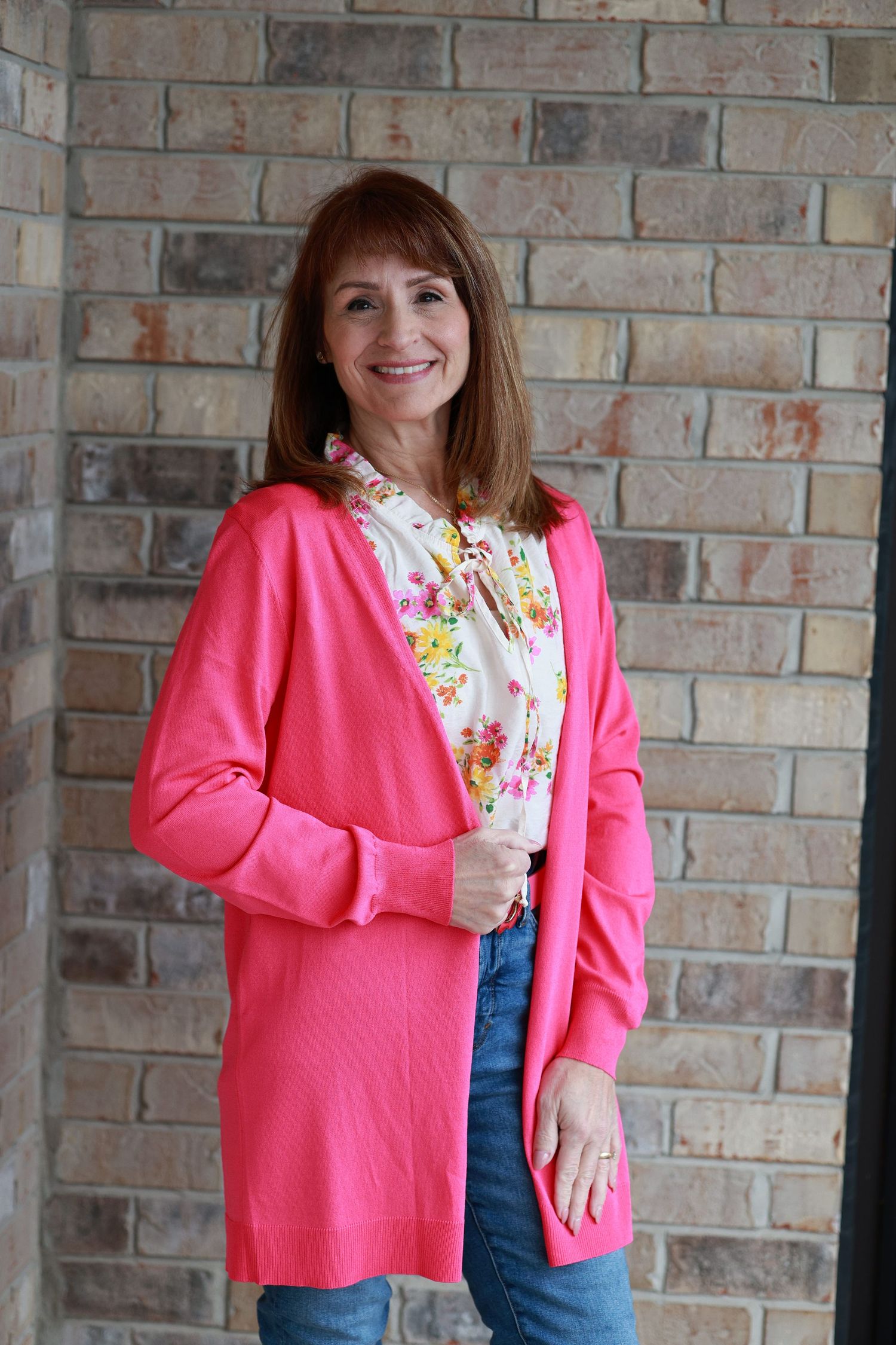 Woman smiling in pink cardigan and floral blouse, jeans, standing against brick wall.