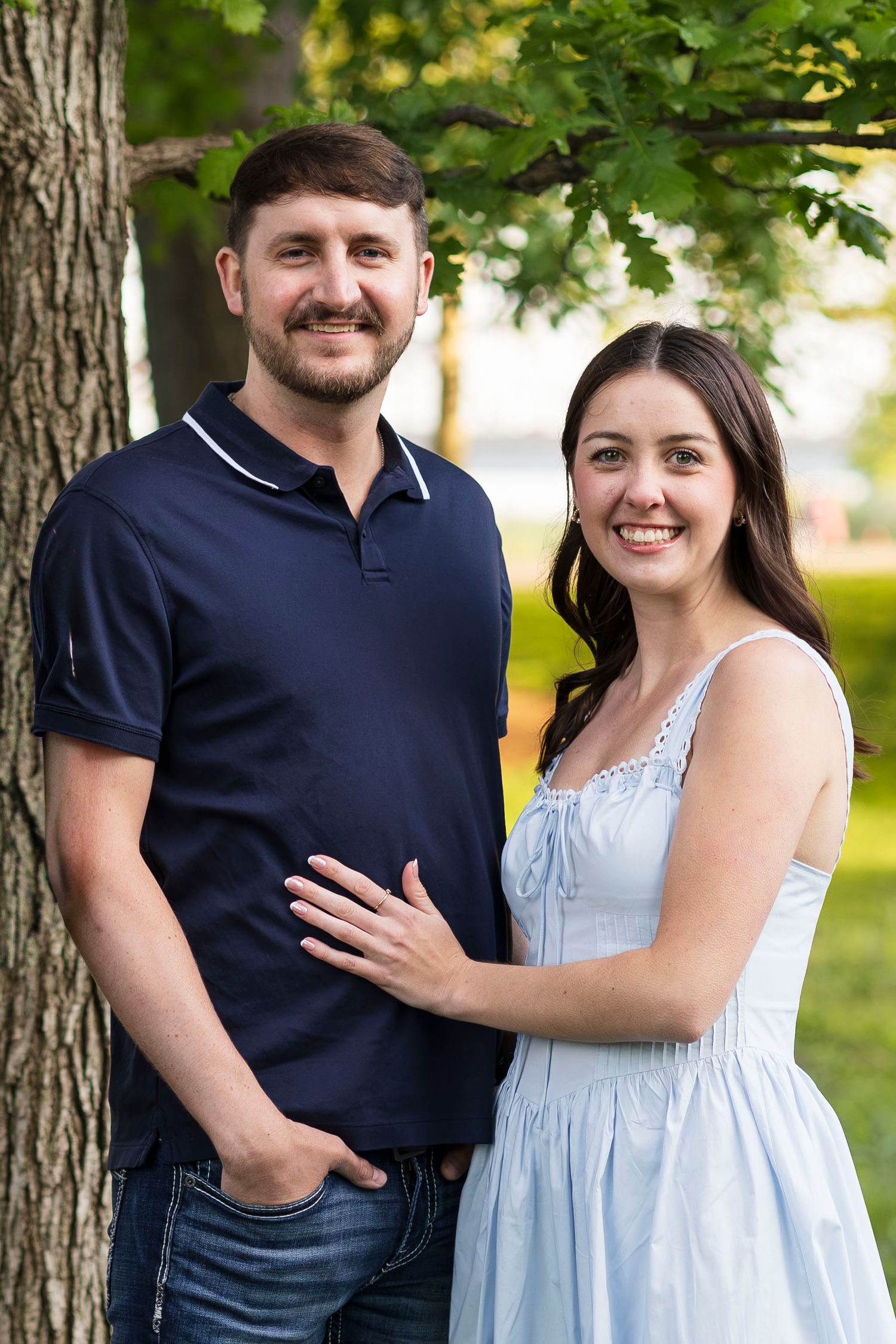 Smiling couple posing outdoors near a tree, man in navy polo and woman in light blue dress.