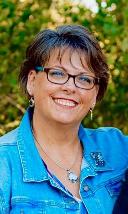 A person in a denim jacket smiling outdoors with natural bokeh background.