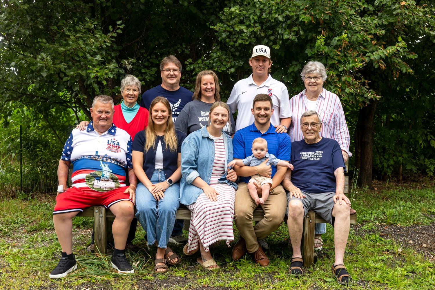 Large multigenerational family group photo outdoors, smiling together in front of lush green trees on a summer day.