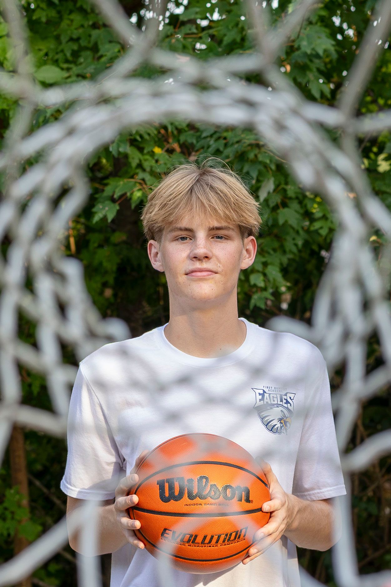 Teen boy holding a Wilson Evolution basketball framed by a hoop net with green trees in the background.
