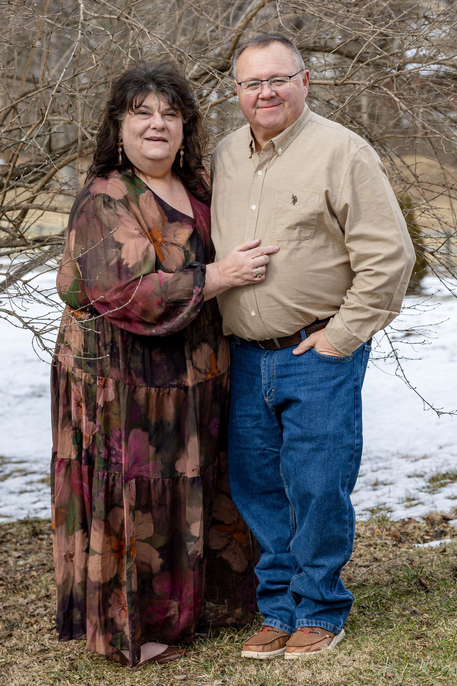 A couple poses together outdoors on a winter day with snow visible in the background.