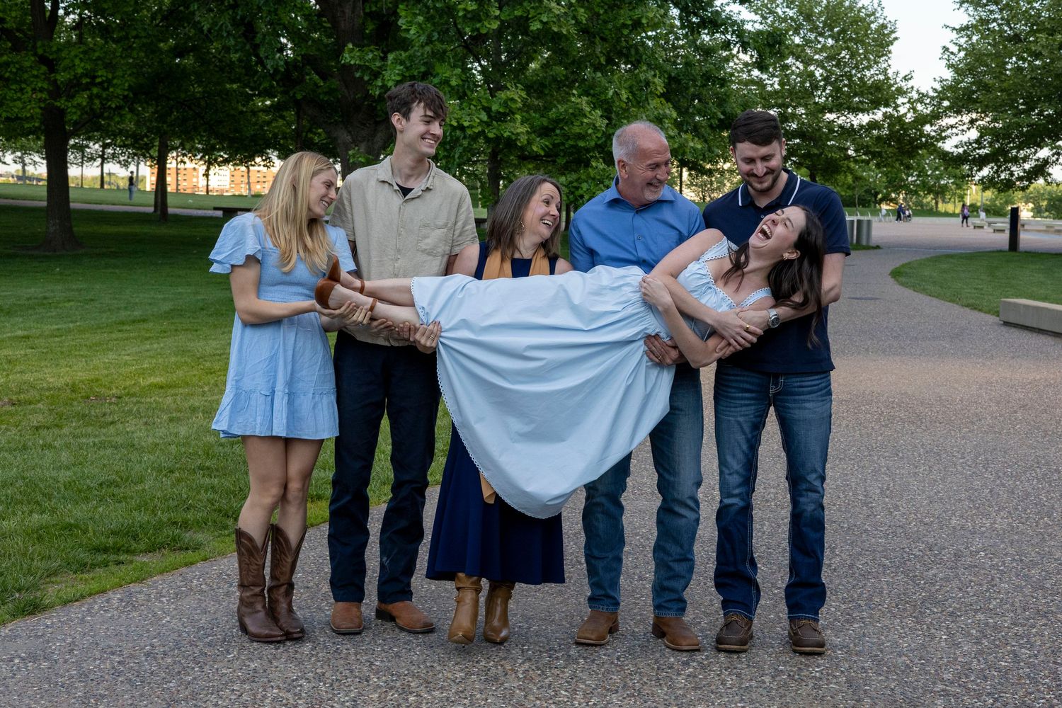 A joyful family group of six adults poses outdoors in a park, with two men carrying a laughing woman horizontally.