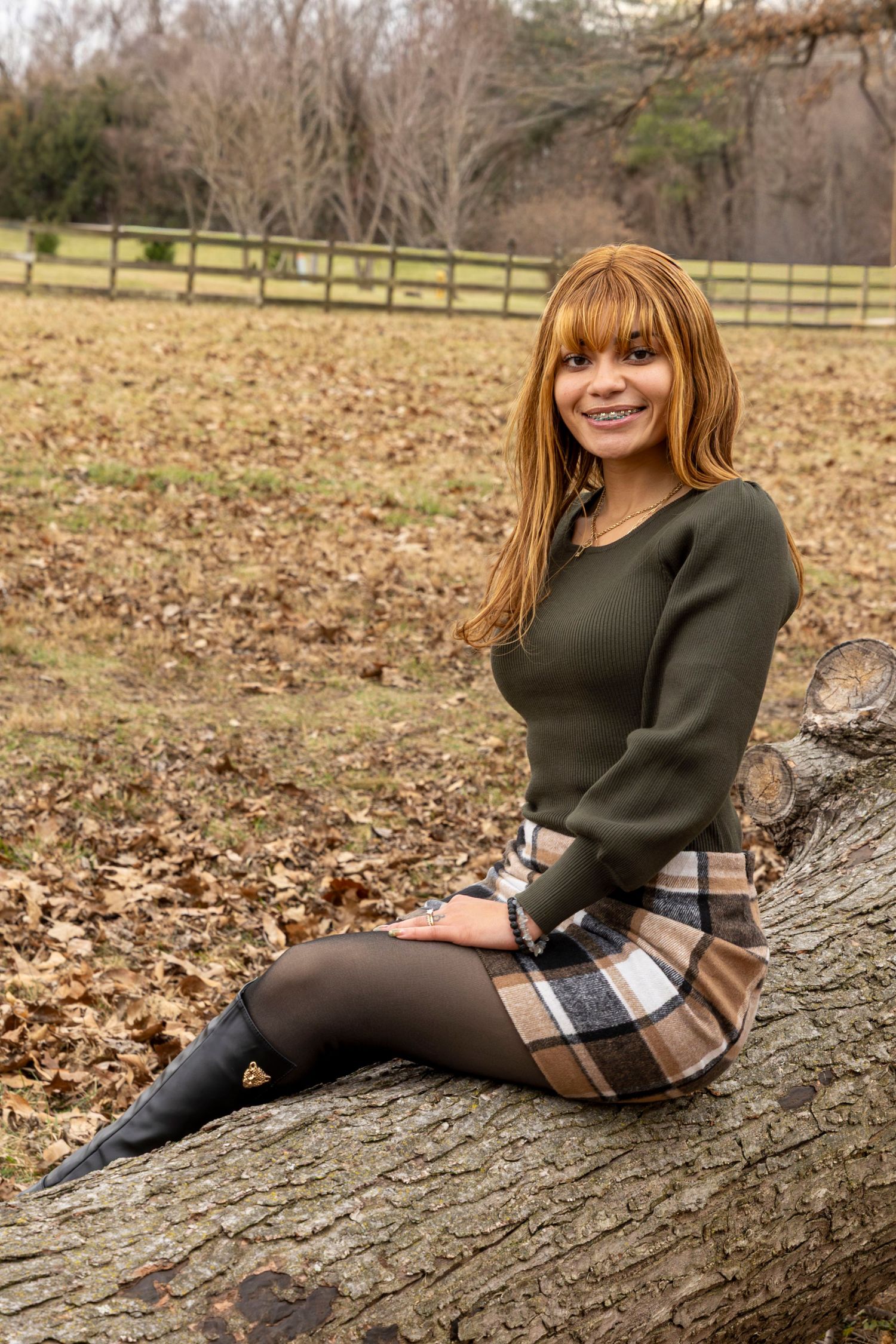 Autumn outdoor portrait with green sweater and plaid skirt sitting on fallen tree trunk in park setting.