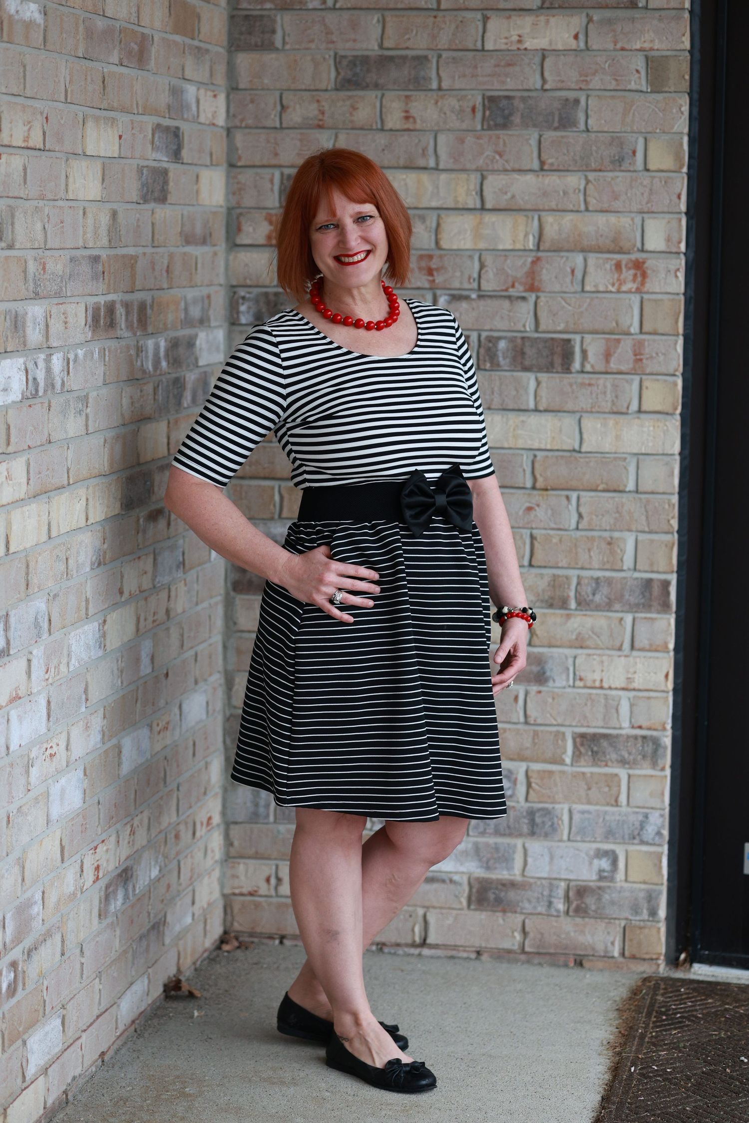 Woman in black and white striped dress with bow belt, red necklace, and flats, posing by brick wall.