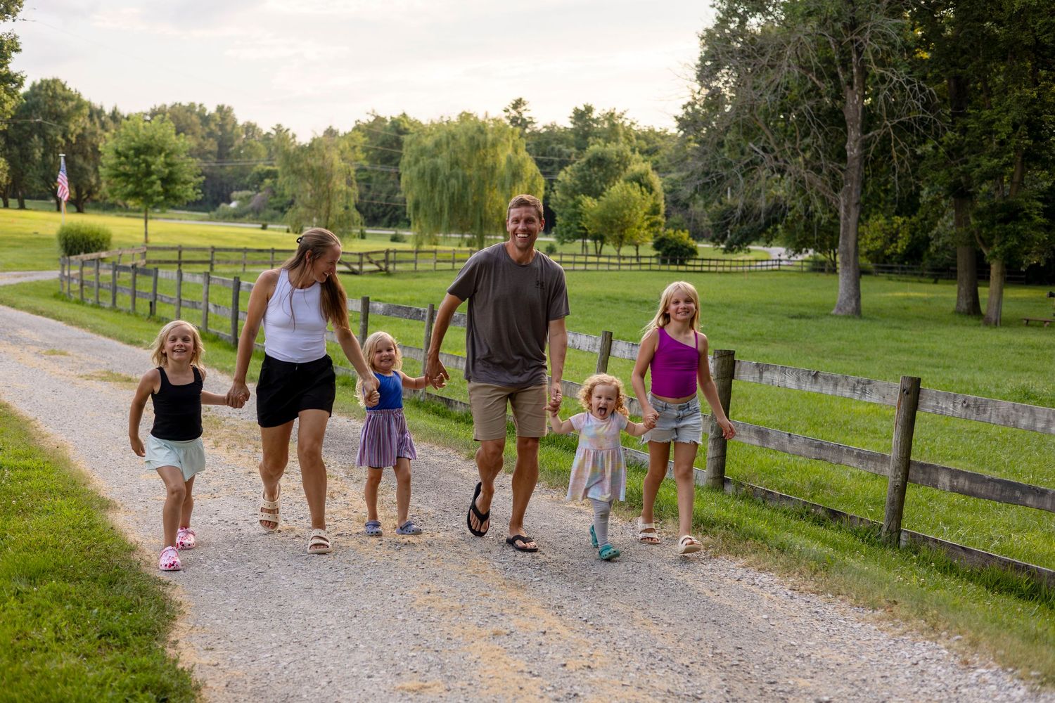 A happy family with four young girls walks hand-in-hand down a scenic gravel path lined with wooden fences.