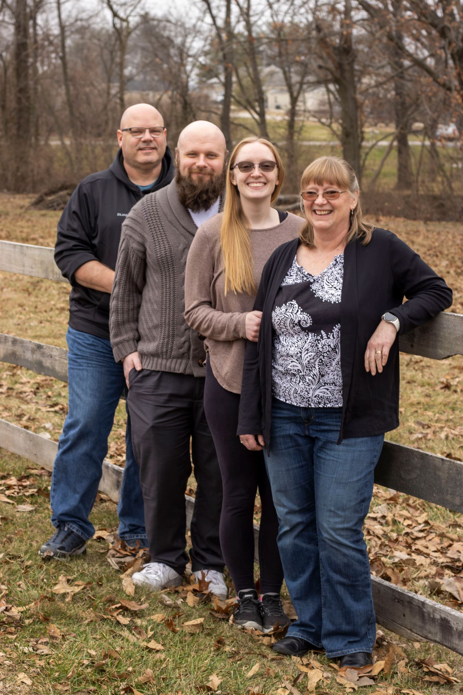 A group of four people standing together outdoors on fallen autumn leaves next to a wooden fence.
