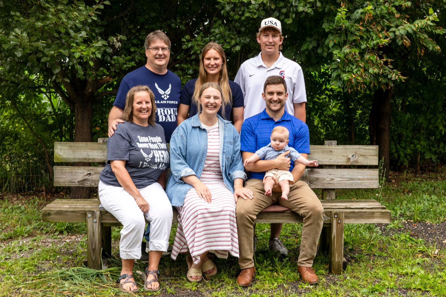 Family portrait of seven people posed on a wooden bench outdoors, including a baby, in a lush green garden setting.
