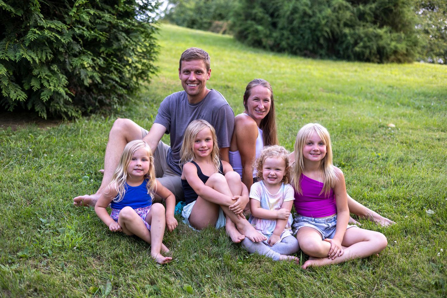 Happy family of two adults and four young girls smiling together on green grass outdoors in summer.