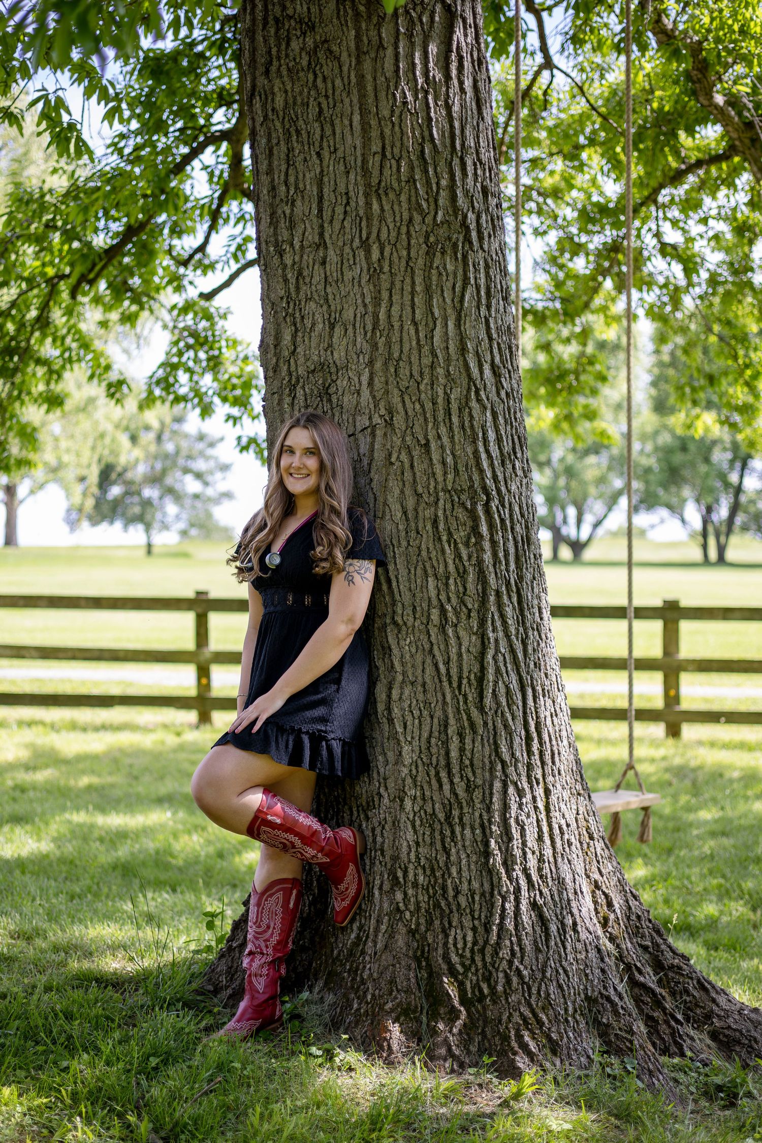Young woman in black dress and red cowboy boots leaning against a large tree with a wooden swing in a green park.