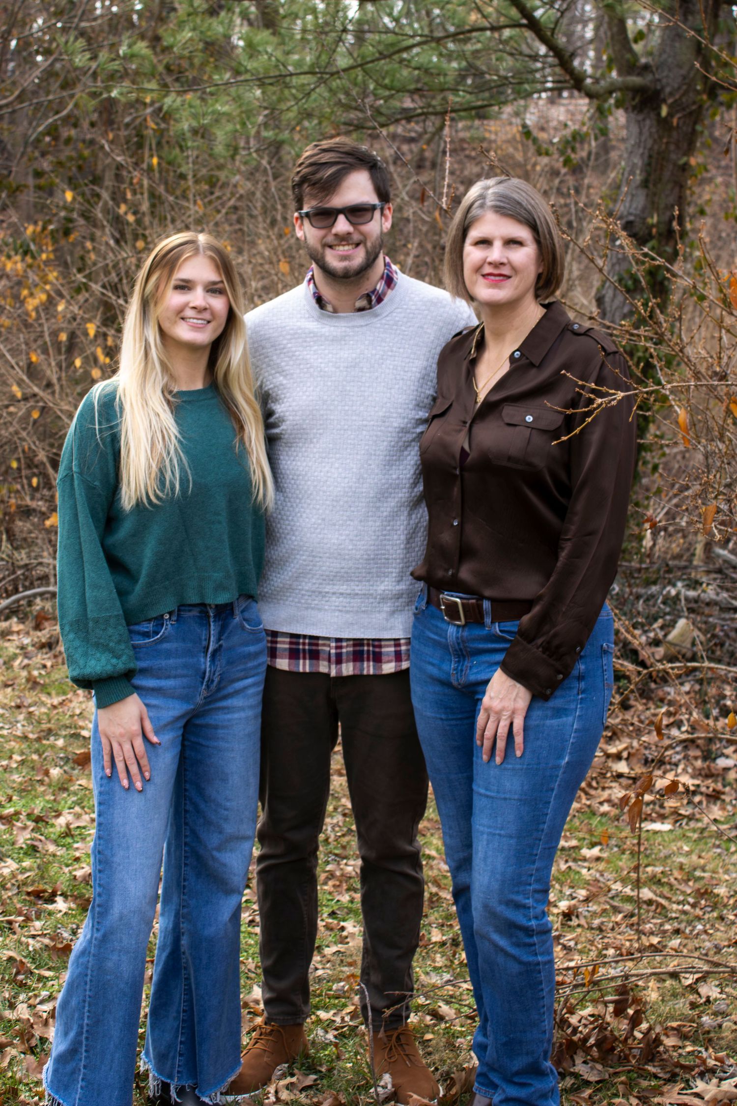Three people stand together outdoors in casual fall attire with jeans and sweaters against an autumn forest backdrop.