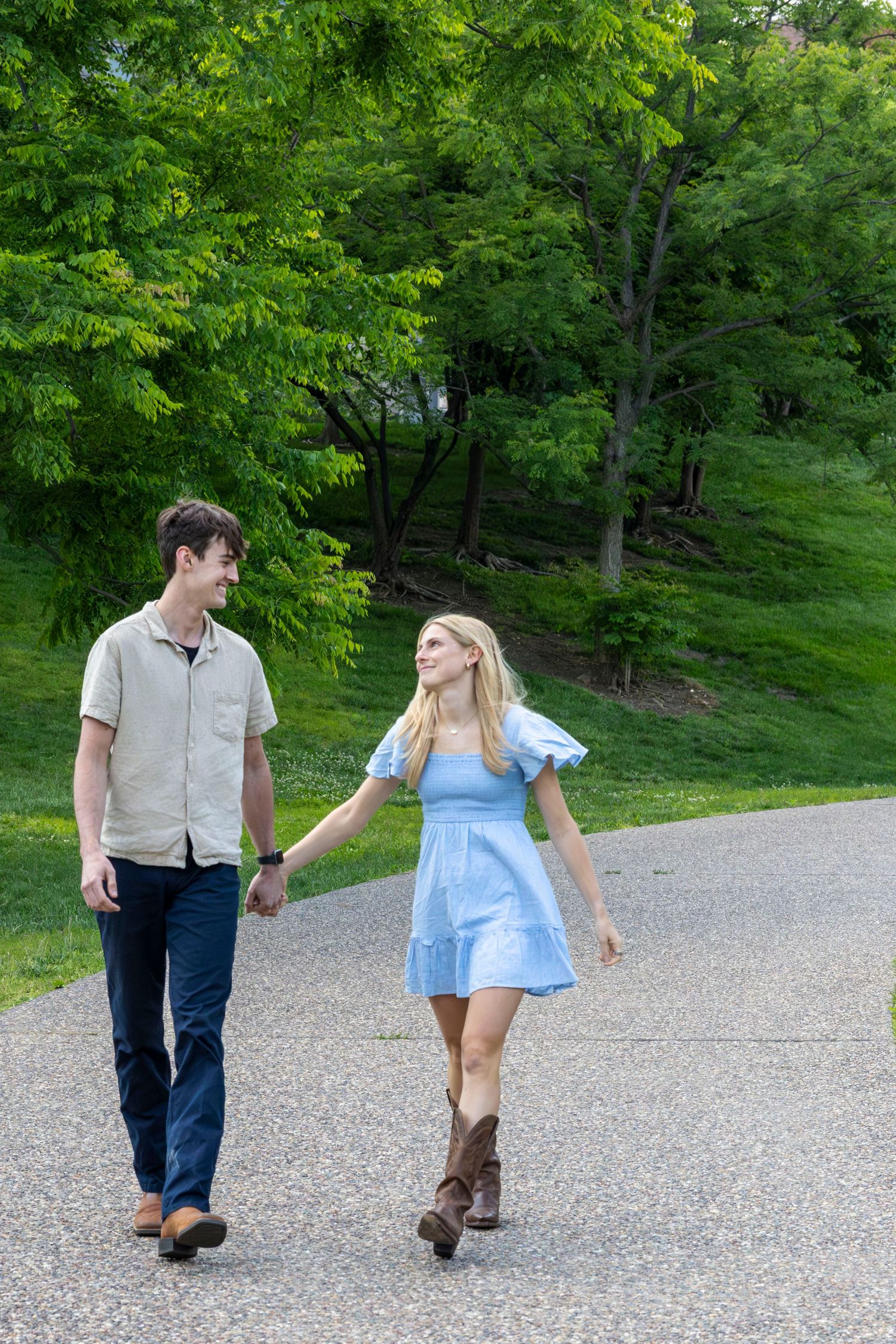 Young couple holding hands walking on a path surrounded by lush green trees on a sunny day.