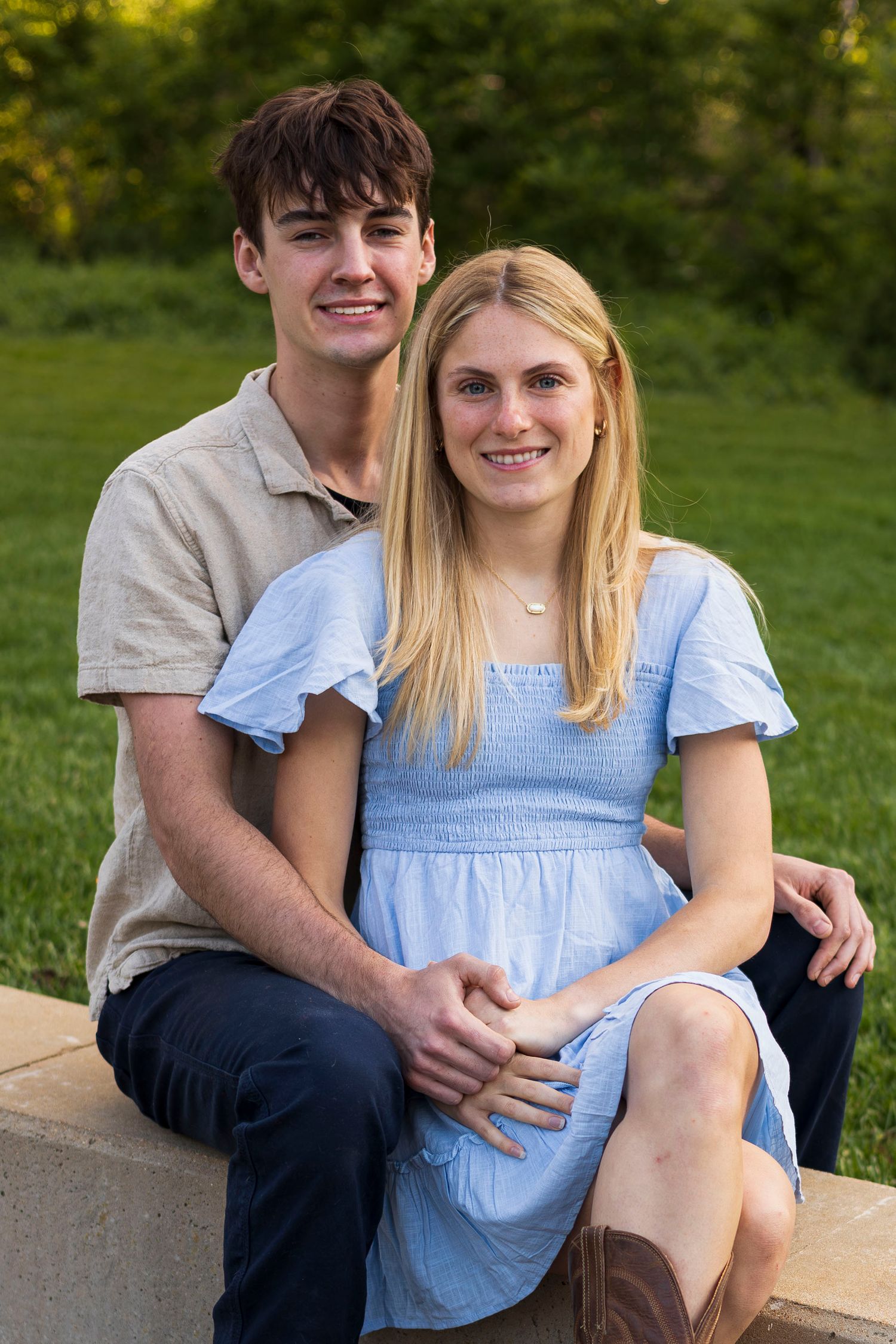 A young couple smiles while sitting together outdoors on stone steps, surrounded by lush green grass.