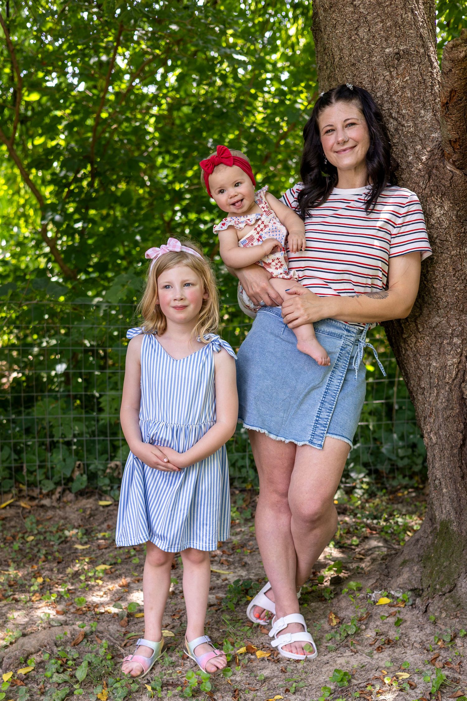 A mother holding a baby with a red bow poses with her young daughter in a blue dress by a tree outdoors.