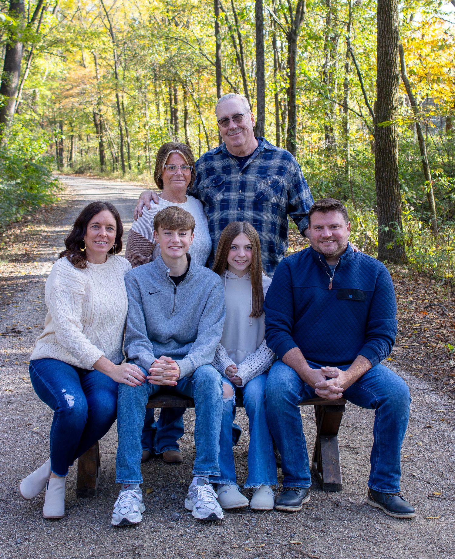 Multi-generational family sitting together on wooden bench in wooded park setting.
