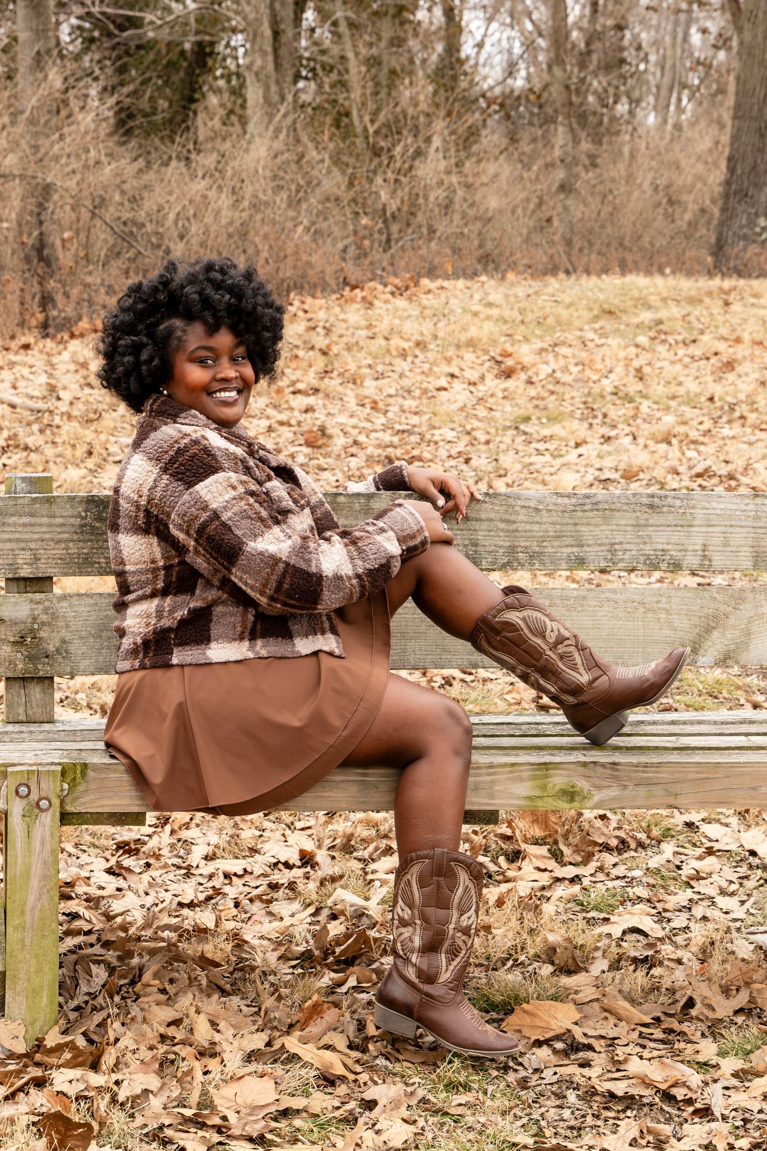 Person in brown boots and plaid shirt poses playfully on wooden bench surrounded by autumn leaves in park setting.