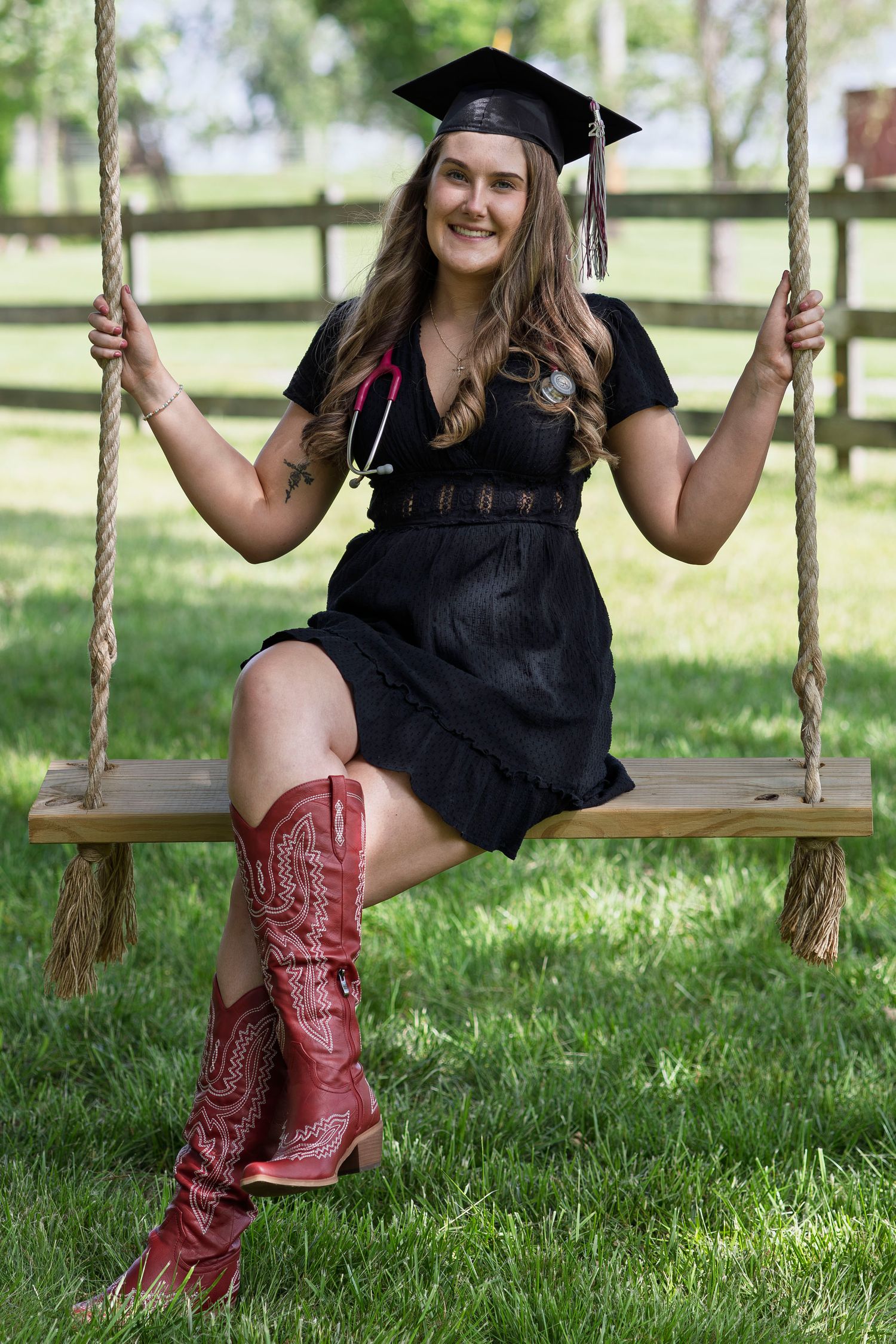A smiling graduate in black dress and red cowboy boots sits on a wooden swing wearing a graduation cap and stethoscope.