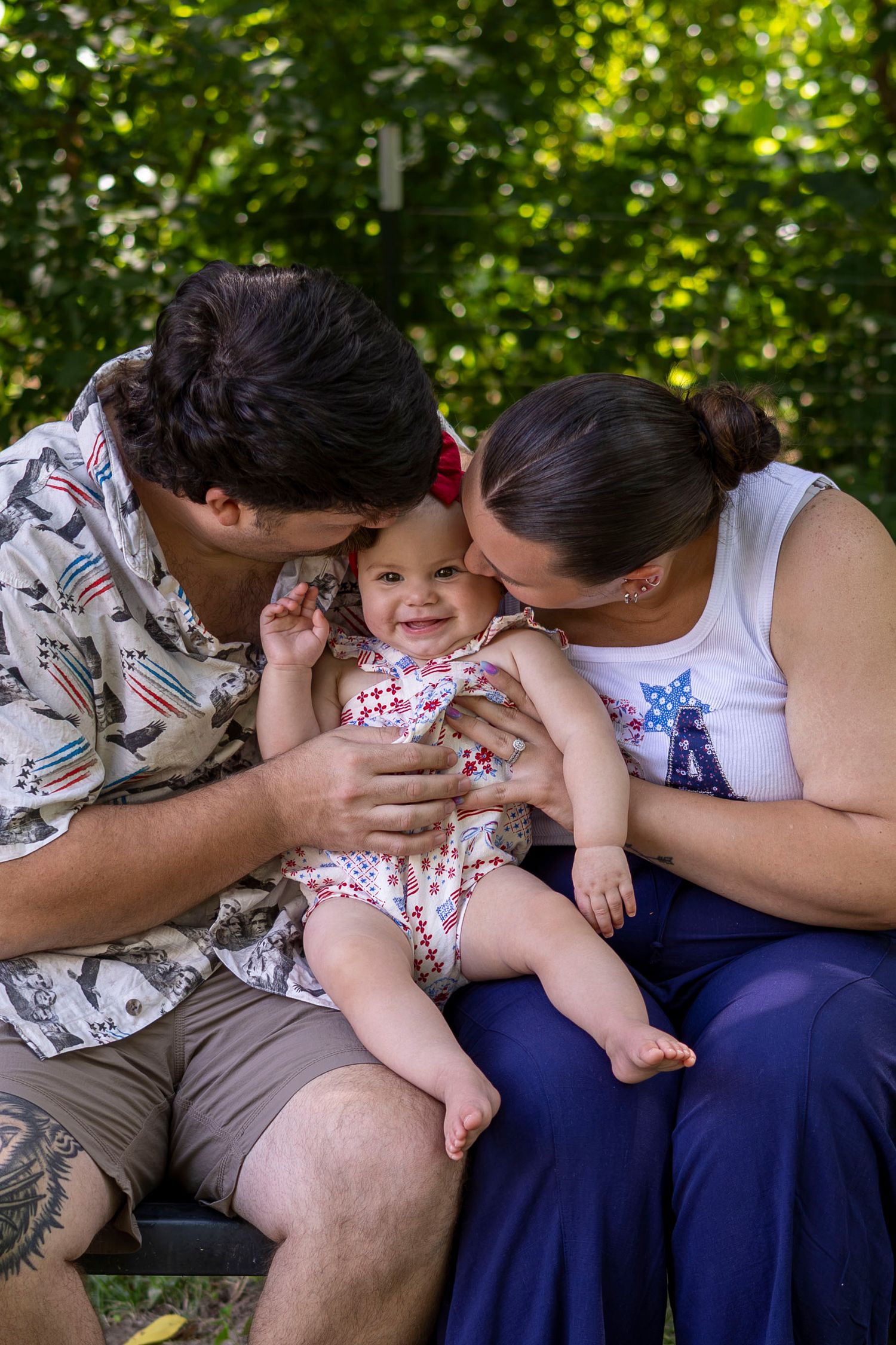 Two parents kissing their smiling baby outdoors, dressed in patriotic outfits among lush green trees.