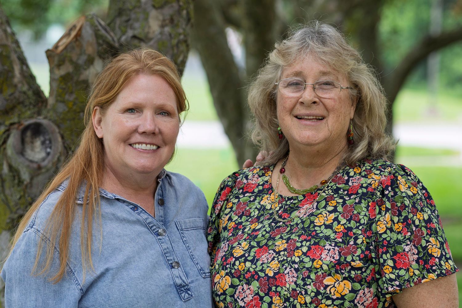 Two smiling women posing outdoors near a large tree on a sunny day, one in denim and one in a floral blouse.