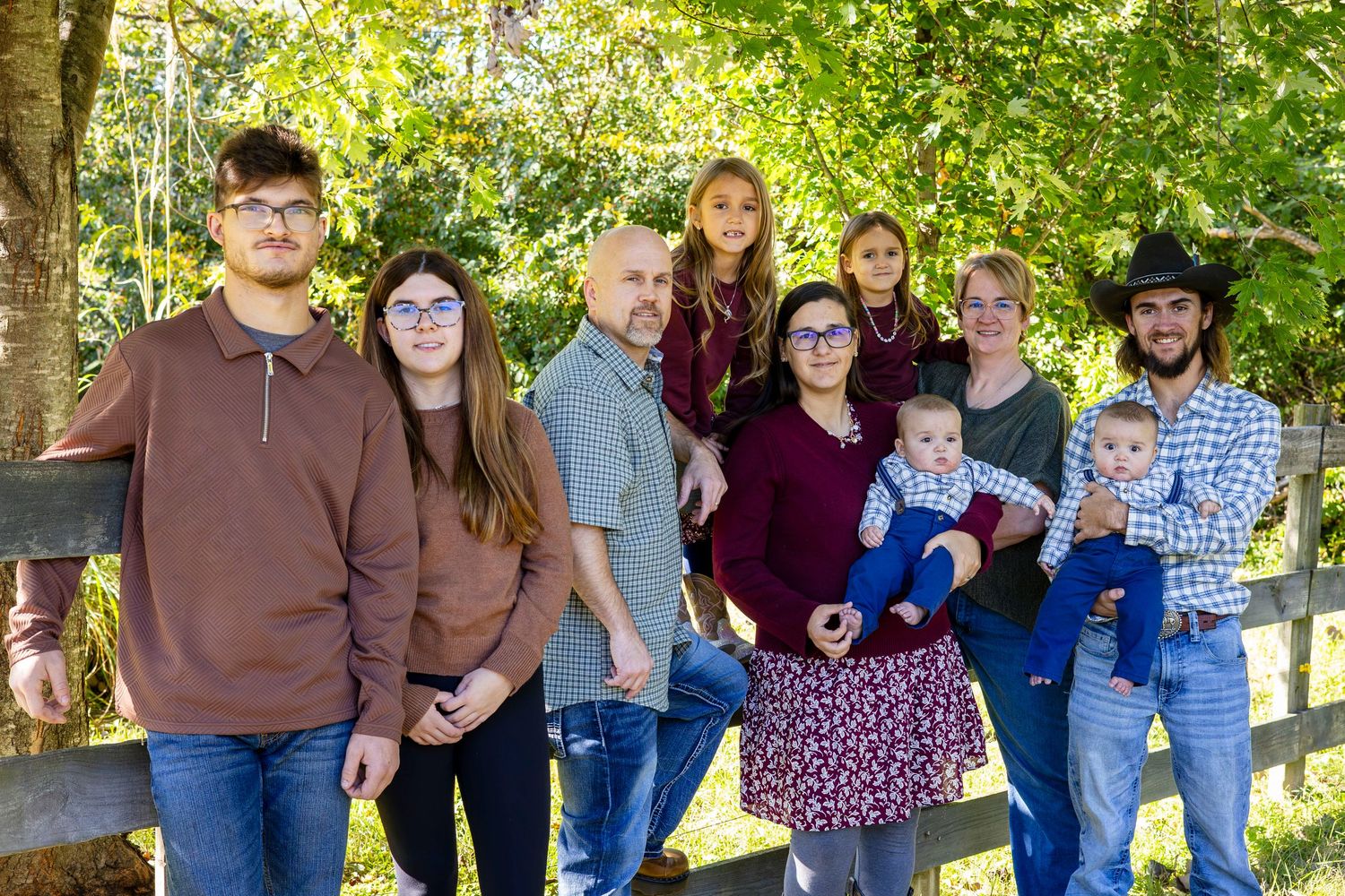 Large family of eleven, including two babies, posing together outdoors beside a wooden fence with green trees in the background.