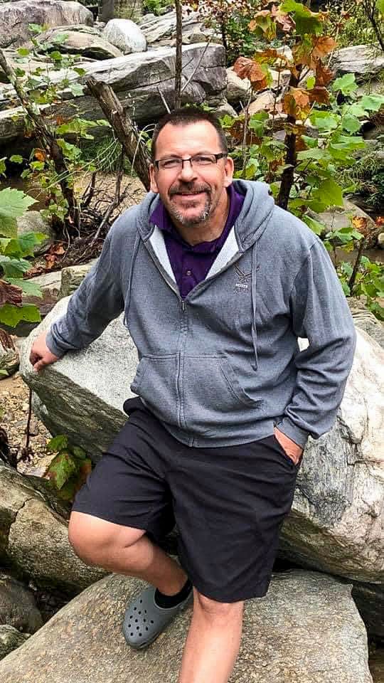 A person in a gray hoodie and shorts sits casually on rocky terrain surrounded by foliage.