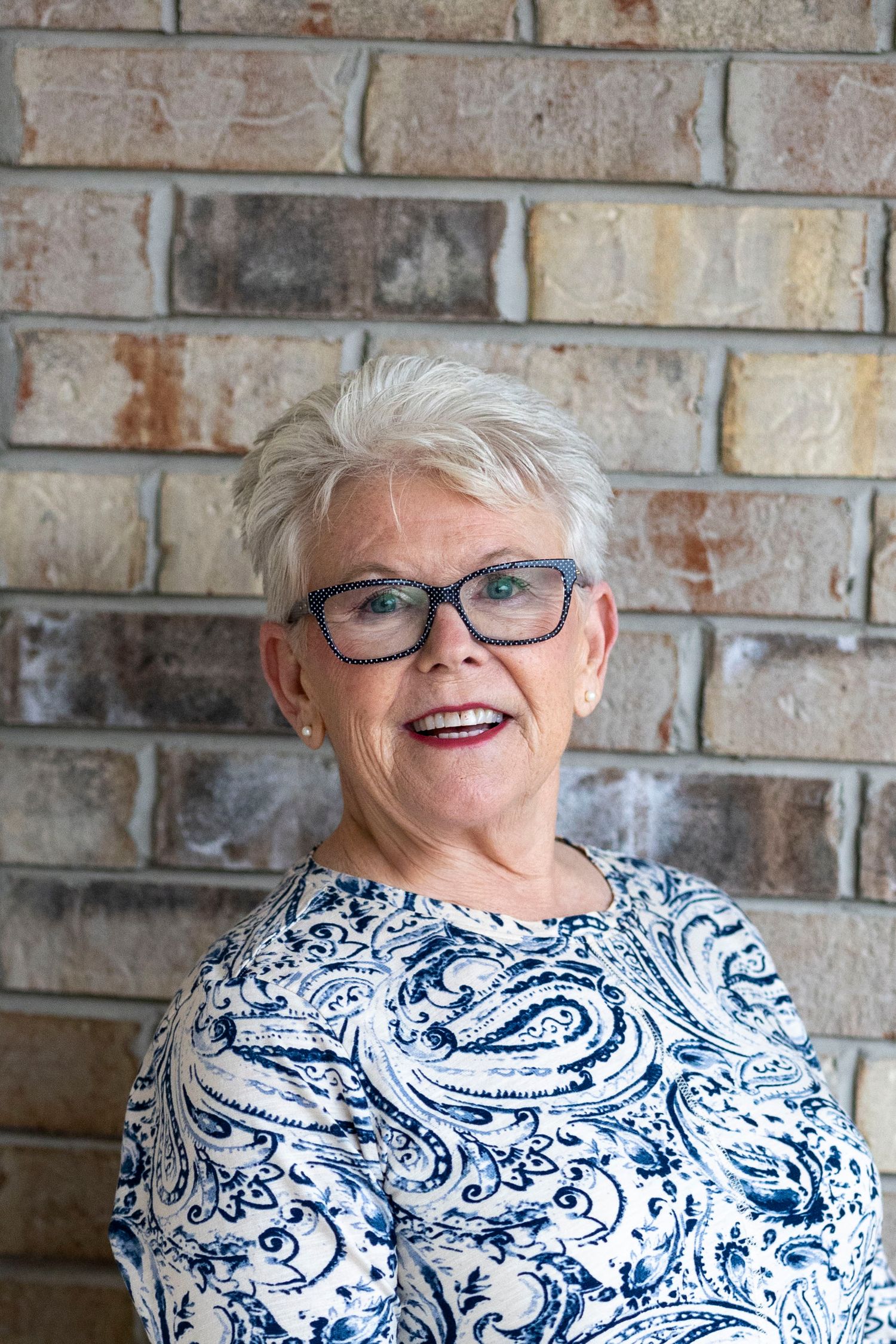 Smiling older woman with short white hair and blue glasses wearing a blue and white patterned blouse against a brick wall.