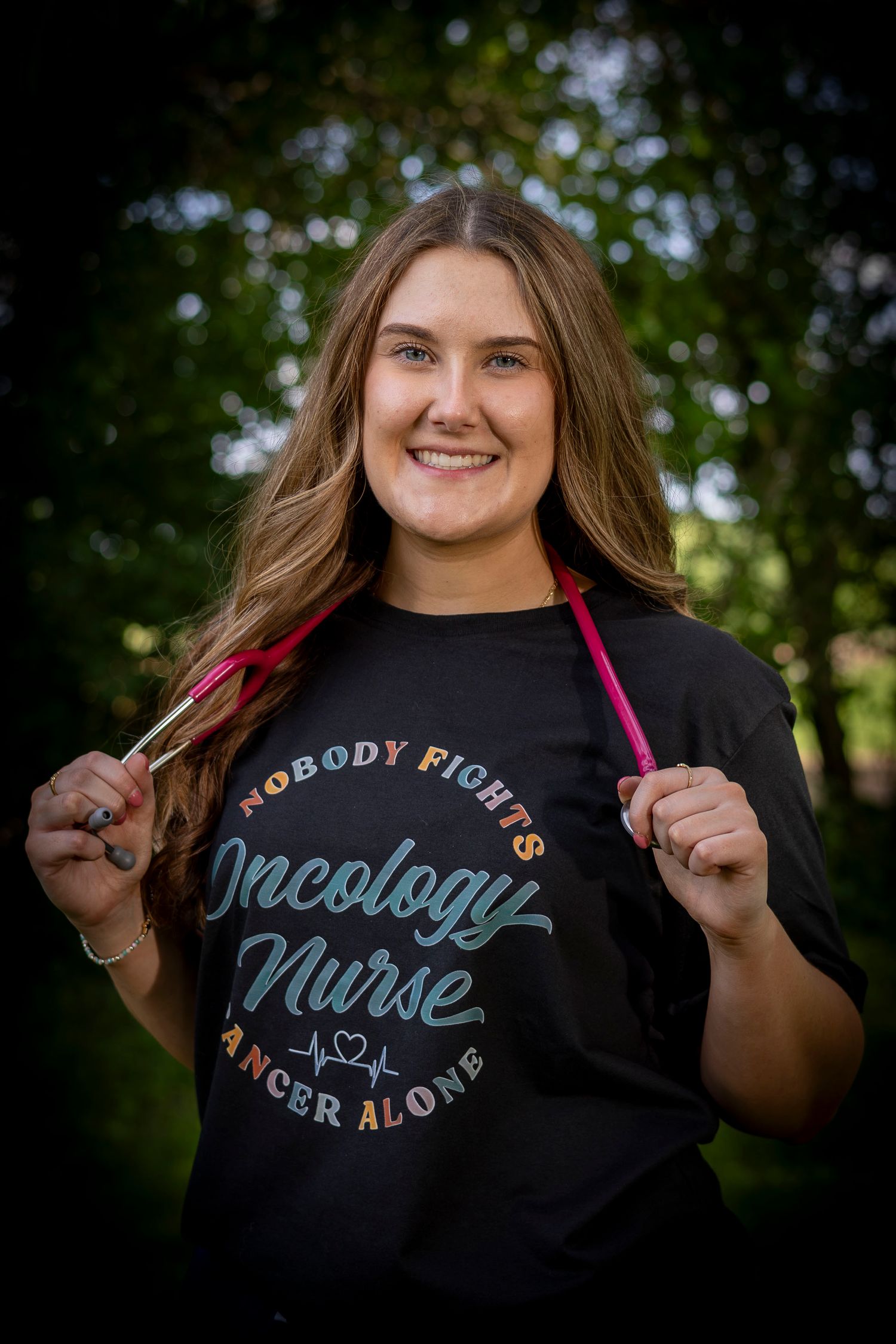 Smiling oncology nurse wearing a black t-shirt that reads Nobody Fights Cancer Alone outdoors.
