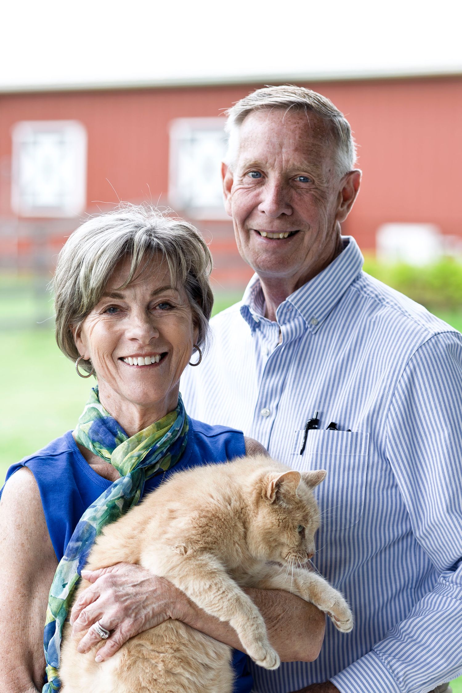 Smiling senior couple holding an orange tabby cat outdoors near a red barn.