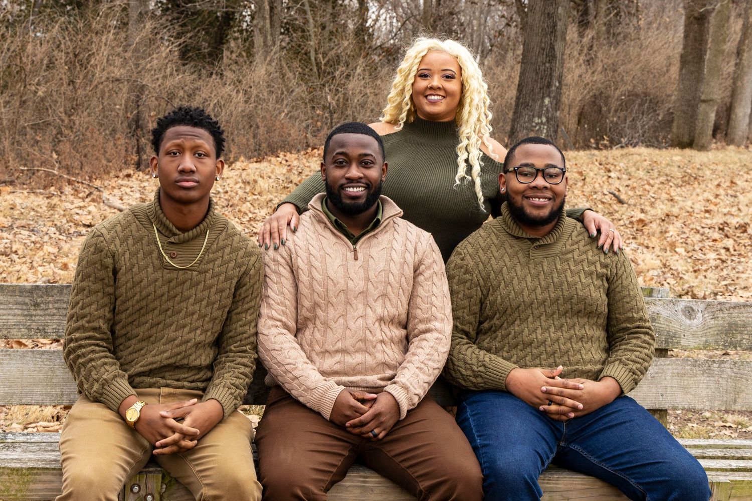 A group of people wearing coordinated olive green and brown outfits sit together outdoors on fallen autumn leaves.