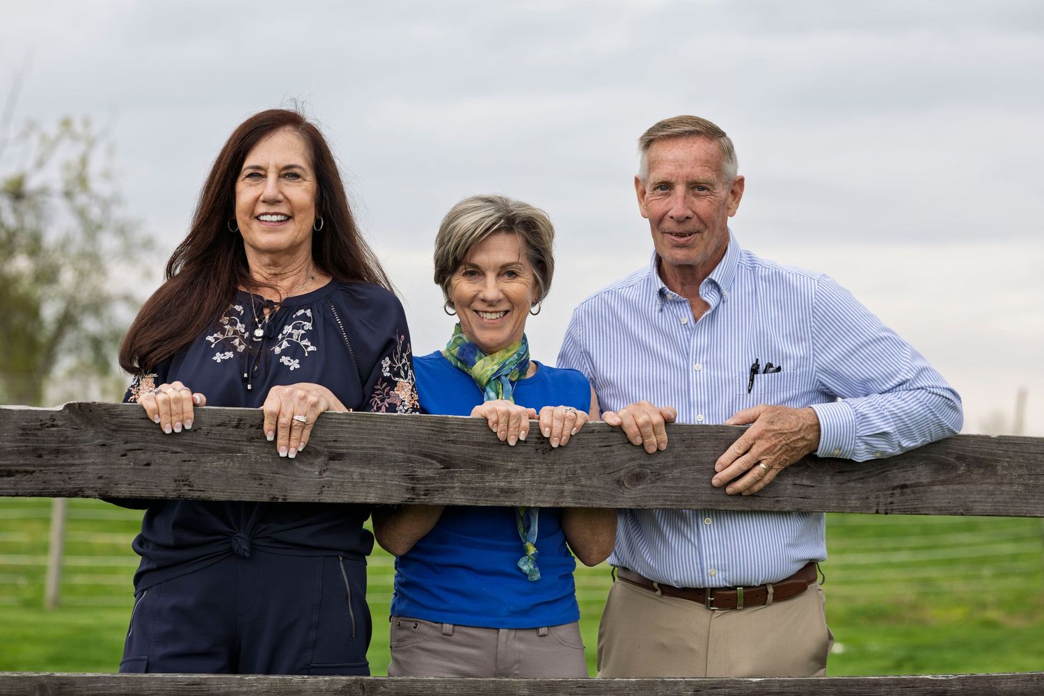 Three smiling adults pose together leaning on a rustic wooden fence outdoors on a sunny day.